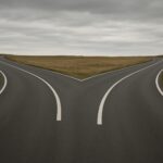 A two-lane road splitting into two directions in a rural landscape under a cloudy sky.