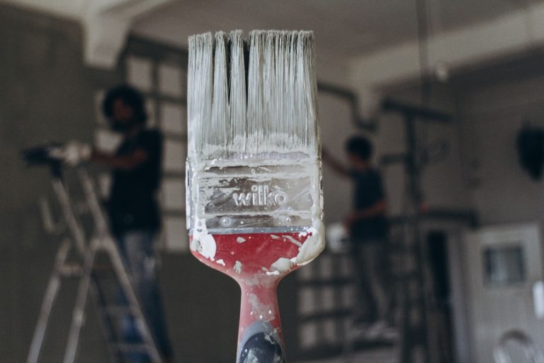 Detailed close-up of a paintbrush with men painting a wall in the blurred background, highlighting renovation work. — Staging